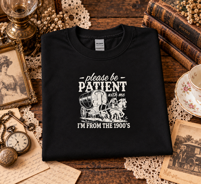 A folded black t-shirt with a white graphic print reading 'please be PATIENT with me I'M FROM THE 1900's' with an illustration of a covered wagon, displayed on a rustic wooden table surrounded by vintage props including books, a teacup, a pocket watch, and lace doilies.