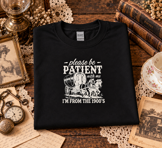 A folded black t-shirt with a white graphic print reading 'please be PATIENT with me I'M FROM THE 1900's' with an illustration of a covered wagon, displayed on a rustic wooden table surrounded by vintage props including books, a teacup, a pocket watch, and lace doilies.