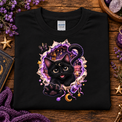A black Gildan Softstyle T-shirt laid flat on a rustic wooden surface, featuring a colorful graphic of a black kitten bursting through a torn purple and gold portal with stars, moons, and a castle. Purple flowers, crystals, a book, and a knitted purple scarf surround the shirt.