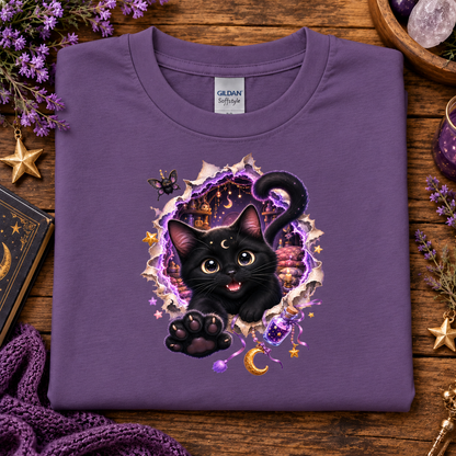A folded heather purple Gildan Softstyle t-shirt with a graphic of a cute black cat bursting from a magical purple and gold portal, displayed on a rustic wooden background with purple-themed decorative props.