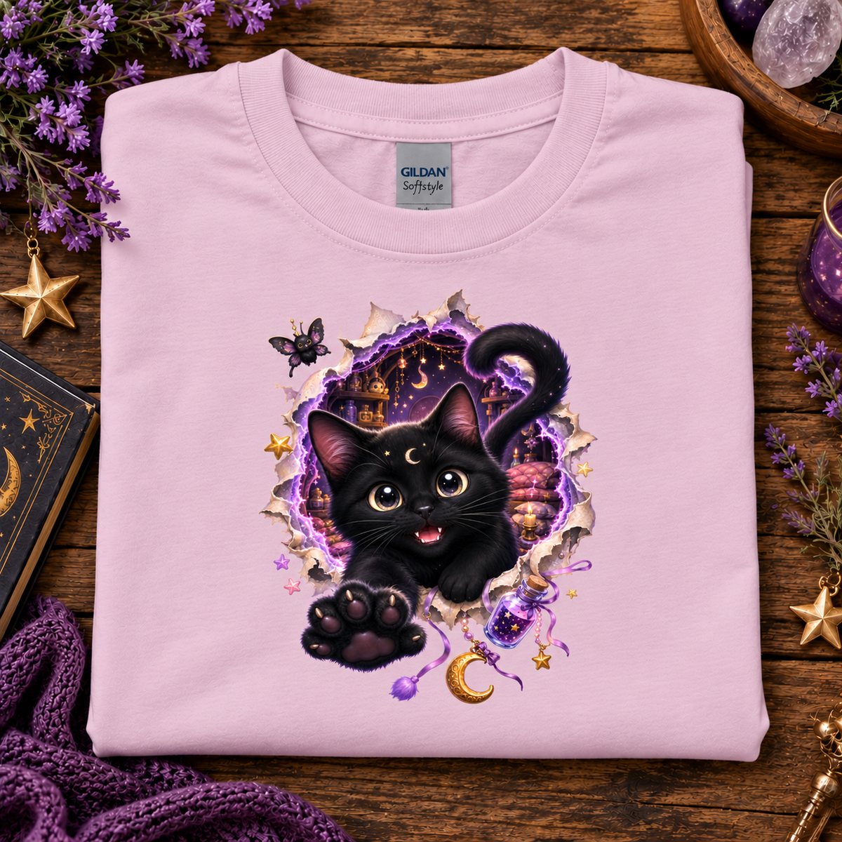 A folded light pink Gildan Softstyle t-shirt with a graphic of a black cat bursting through a torn purple and gold portal, displayed on a rustic wooden background with decorative purple flowers, a book, and a purple knit blanket.