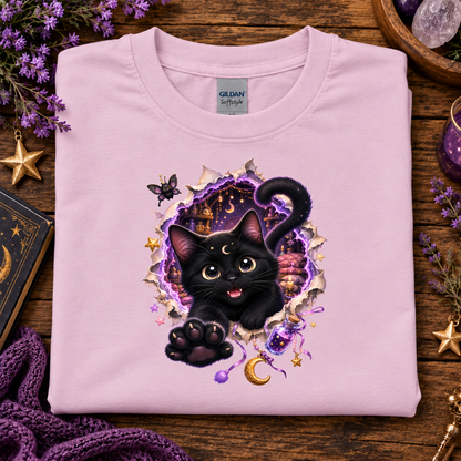 A folded light pink Gildan Softstyle t-shirt with a graphic of a black cat bursting through a torn purple and gold portal, displayed on a rustic wooden background with decorative purple flowers, a book, and a purple knit blanket.