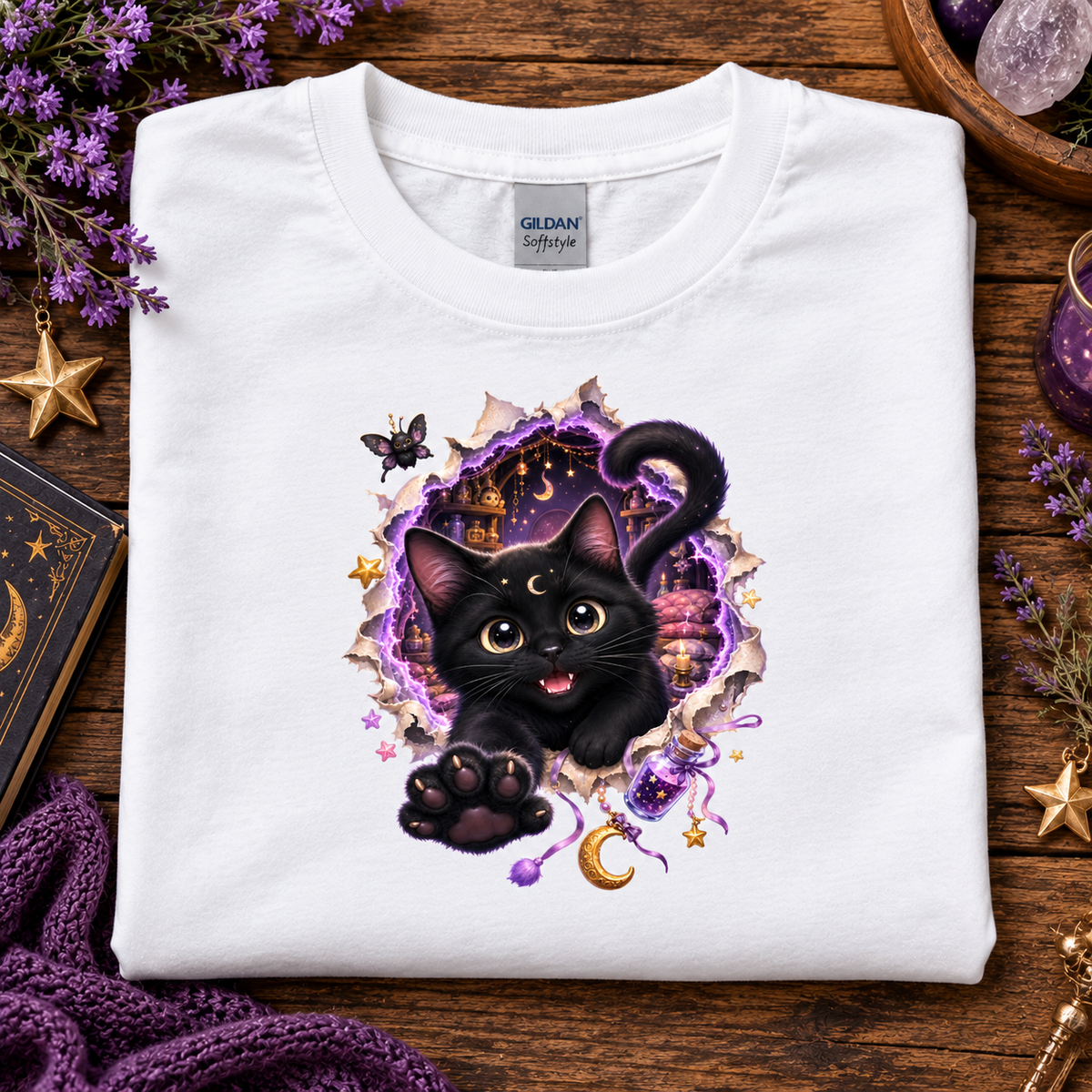 A folded white Gildan Softstyle T-shirt with a graphic of a black cat bursting through torn purple and gold paper, surrounded by stars, a crescent moon, and magical elements, displayed on a wooden surface with decorative props.
