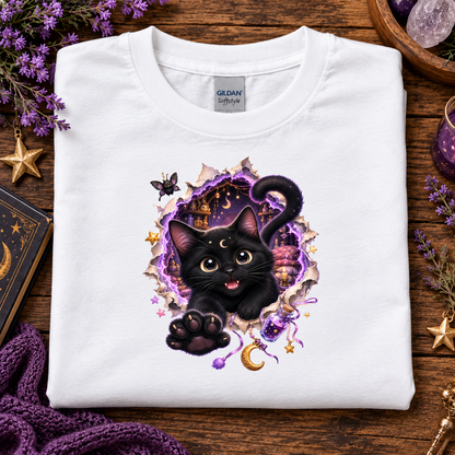 A folded white Gildan Softstyle T-shirt with a graphic of a black cat bursting through torn purple and gold paper, surrounded by stars, a crescent moon, and magical elements, displayed on a wooden surface with decorative props.