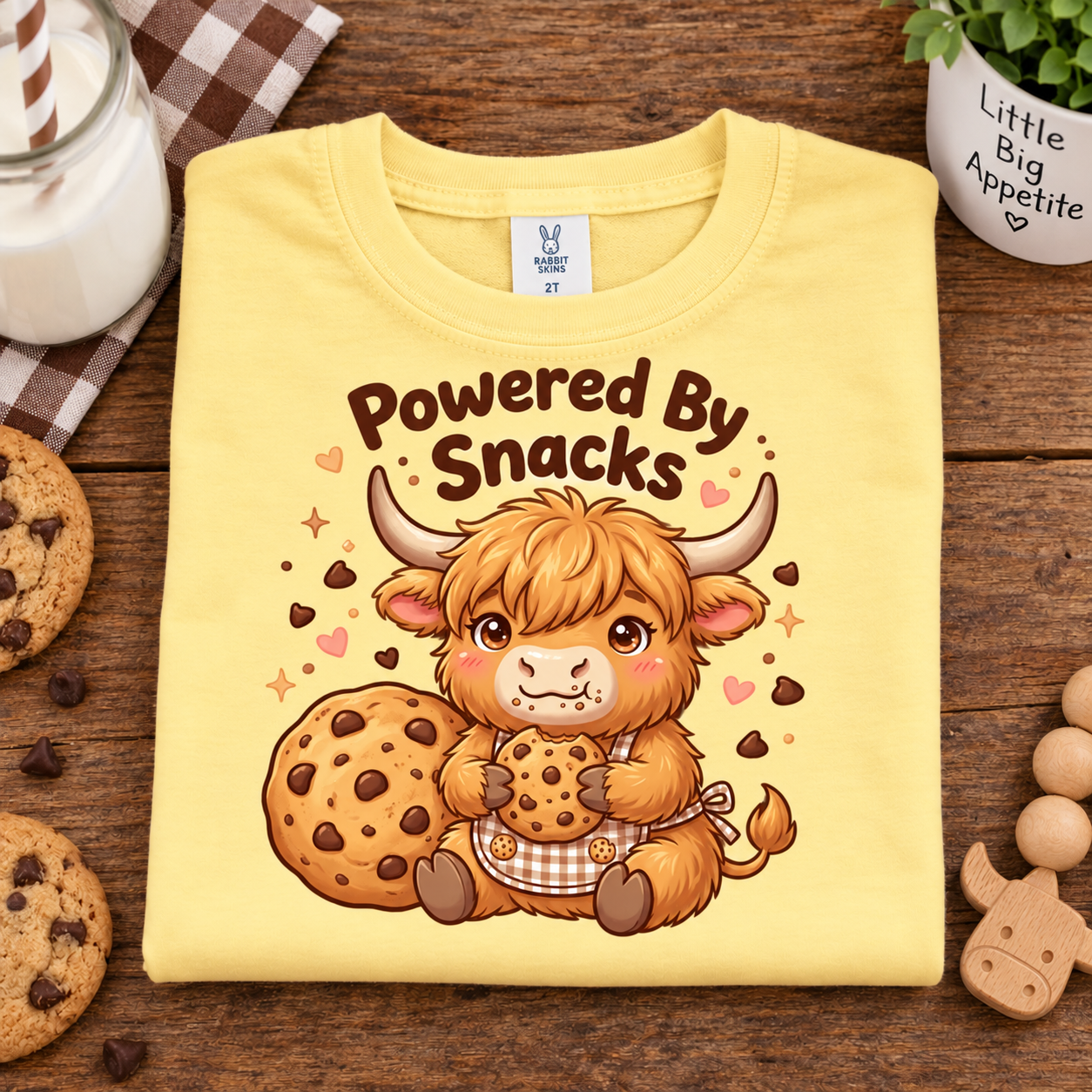 A folded butter colored T-shirt with a cartoon Highland cow holding cookies and the text 'Powered By Snacks' on the front, styled on a wooden surface with cookies, a mug, and a small plant.