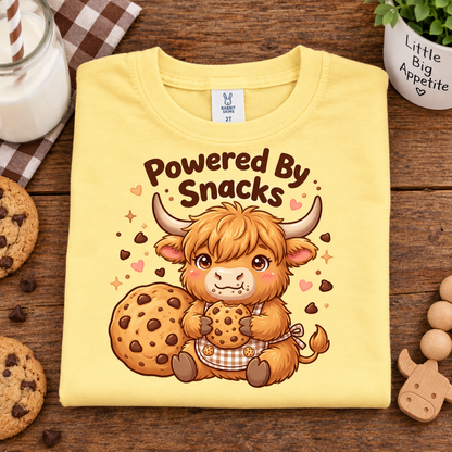 A folded butter colored T-shirt with a cartoon Highland cow holding cookies and the text 'Powered By Snacks' on the front, styled on a wooden surface with cookies, a mug, and a small plant.