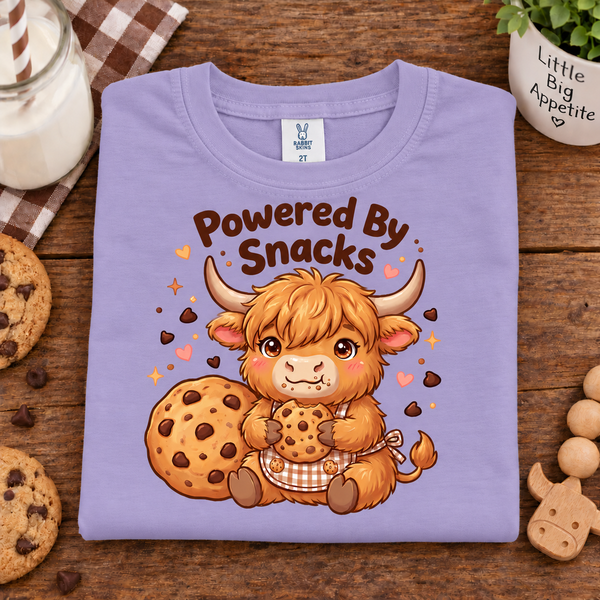 A folded lavender crewneck t-shirt with a cartoon Highland cow holding cookies and a chocolate chip cookie, with the text 'Powered By Snacks' above it. The shirt is displayed on a wooden table surrounded by cookies, a glass of milk, and a small plant.