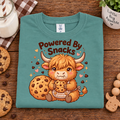 A apple colored folded t-shirt with a cartoon Highland cow holding cookies and the text 'Powered By Snacks' on the front, styled on a wooden table with cookies, a candle, and a small plant.