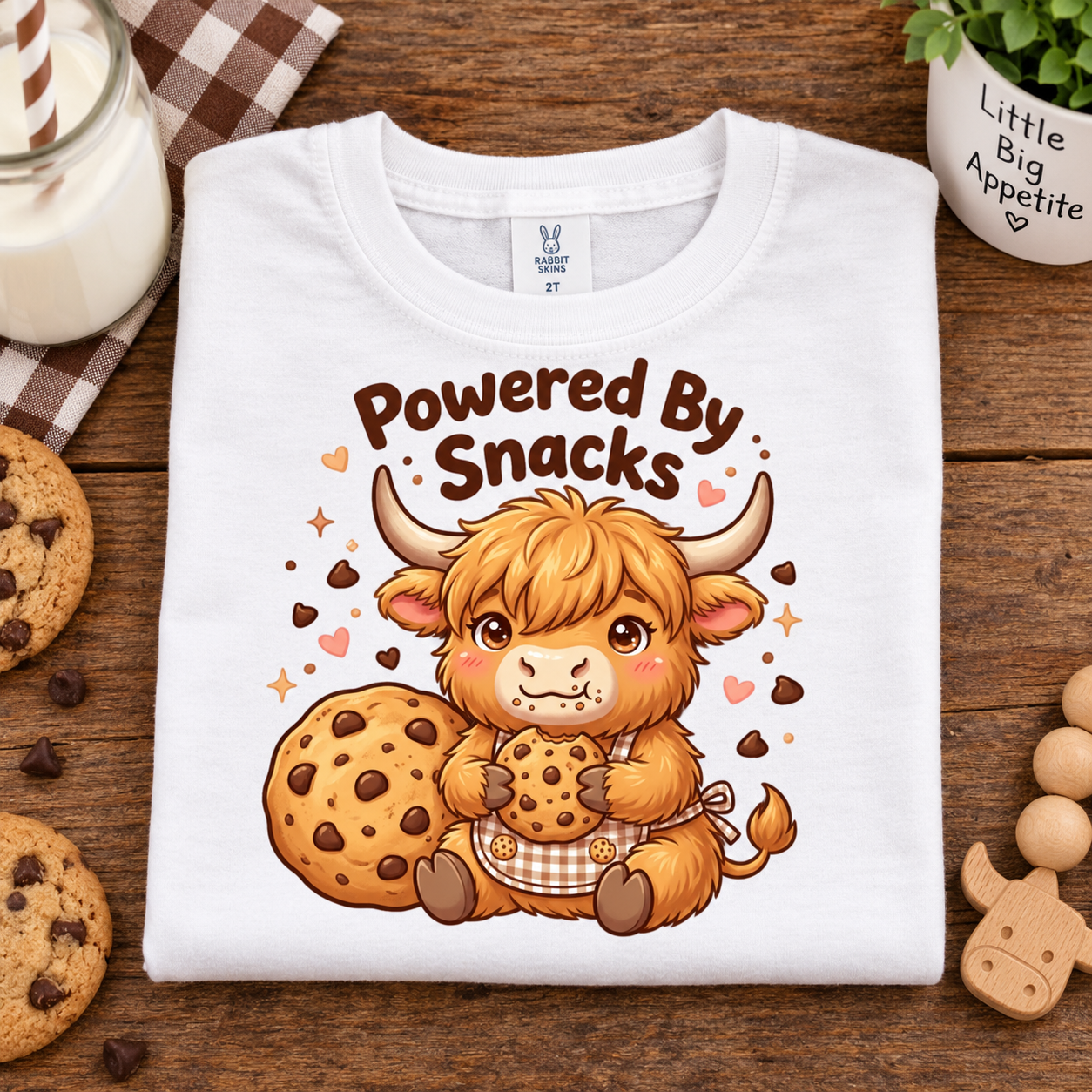 A folded white t-shirt with a cute cartoon Highland cow graphic holding cookies and a cookie, with the text 'Powered By Snacks'. The t-shirt is displayed in a flat-lay setting on a wooden table surrounded by cookies, a glass of milk, and a small plant.