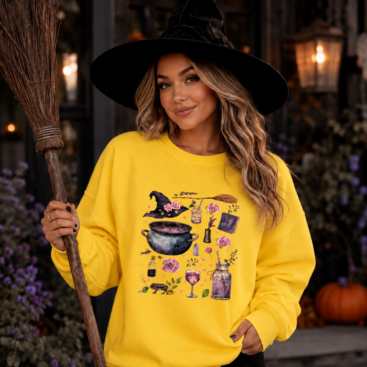 A woman wearing a bright yellow long-sleeve sweatshirt with a witch-themed graphic print, holding a broomstick in a Halloween-themed setting. The graphic features a cauldron, potion bottle, rose, and broom.