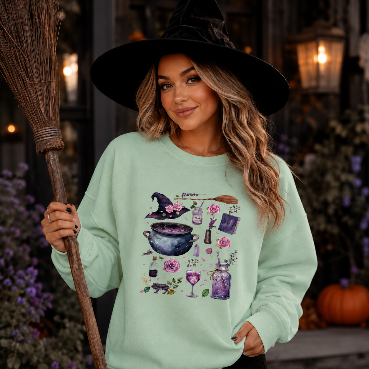 A woman wearing a black witch hat and a mint green graphic sweatshirt, holding a broomstick in a Halloween-themed setting with pumpkins and purple flowers.
