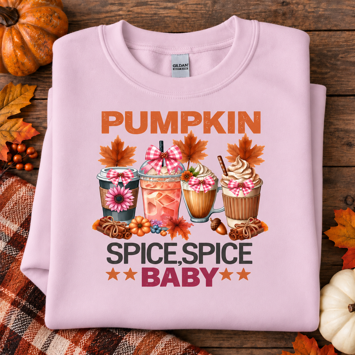 A folded light pink crewneck sweatshirt with a colorful autumn-themed graphic reading 'PUMPKIN SPICE, SPICE BABY' and featuring illustrated drinks, pumpkins, leaves, and cinnamon sticks, displayed on a rustic wood background with fall leaves and a plaid blanket.