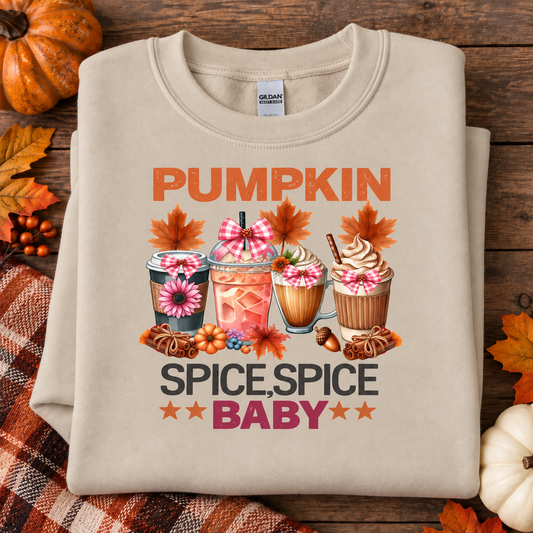 A folded sand crewneck sweatshirt with a colorful autumn-themed graphic reading 'PUMPKIN SPICE, SPICE BABY' in orange, pink, and black text, surrounded by illustrated drinks, flowers, leaves, and candy. The sweatshirt is styled on a rustic wooden surface with fall leaves, a pumpkin, and a plaid blanket.