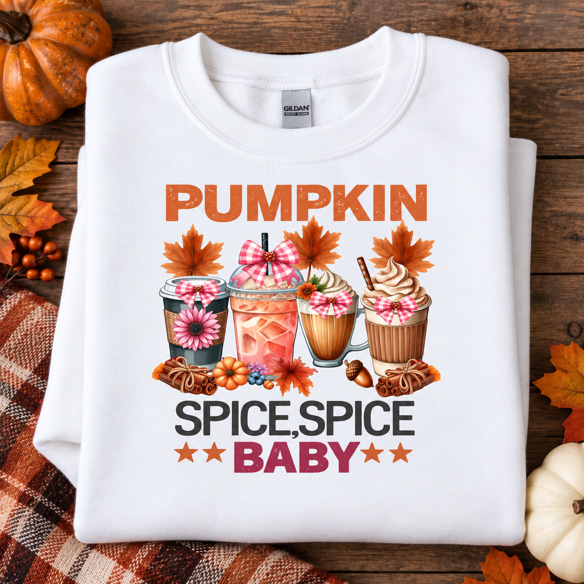 A folded white Gildan sweatshirt with a colorful autumn graphic reading 'PUMPKIN SPICE, SPICE BABY,' styled on a rustic wood surface with pumpkins, fall leaves, and a plaid cloth.