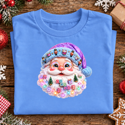 A folded carolina blue t-shirt with a large, detailed 3D Santa Claus face graphic on the chest, displayed on a rustic wooden background decorated with Christmas ornaments, pine branches, and a gingerbread cookie.