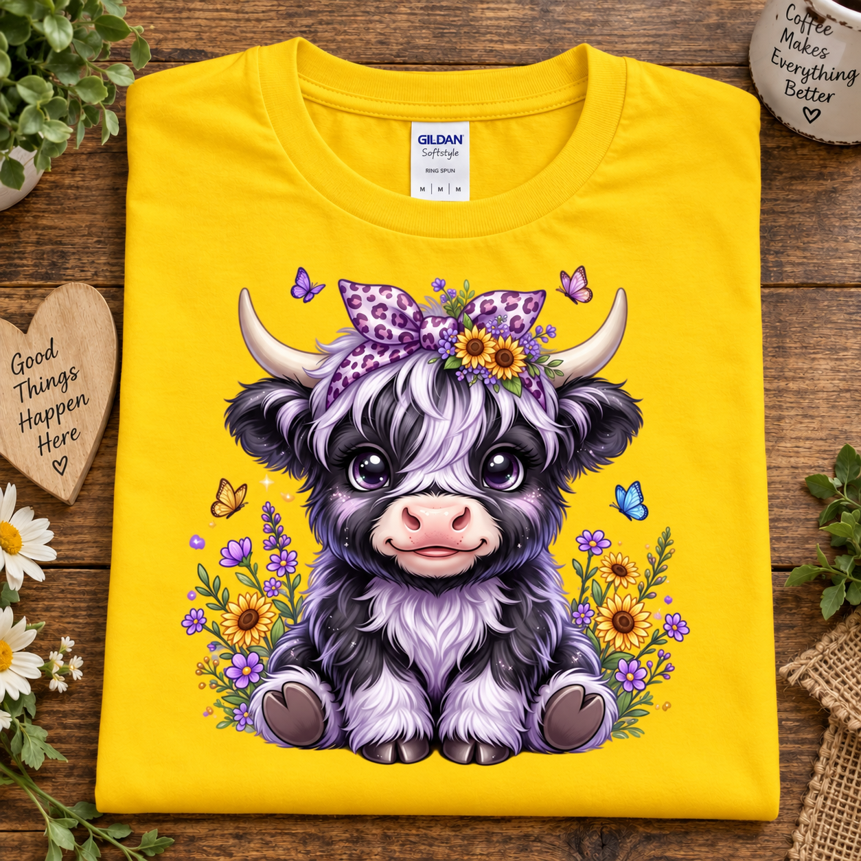 A folded daisy colored Gildan Comfort T-shirt is displayed on a rustic wooden surface. The shirt features a large, colorful cartoon graphic of a cute black and white highland cow wearing a purple bow and flower crown, surrounded by wildflowers and butterflies.