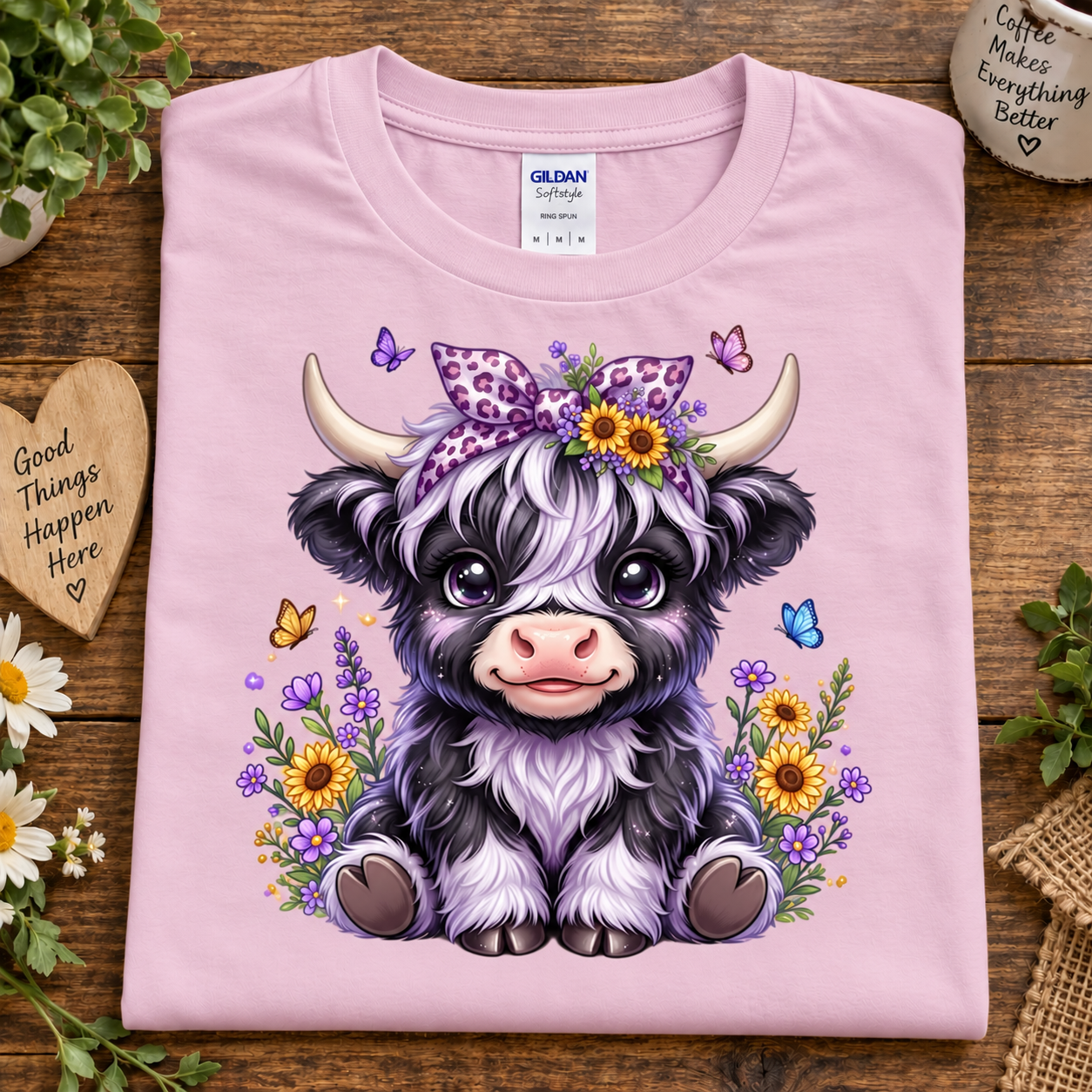 A folded light pink Gildan cotton t-shirt with a cute cartoon illustration of a black and white highland cow wearing a purple leopard-print bow and surrounded by yellow and purple flowers, displayed on a rustic wooden background with decorative props.