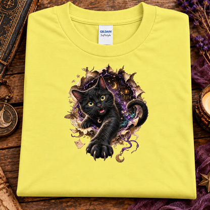 A folded cornsilk yellow Gildan Softeagle t-shirt featuring a graphic of a black cat bursting through a magical, purple and gold portal, displayed on a rustic wooden background with decorative props.