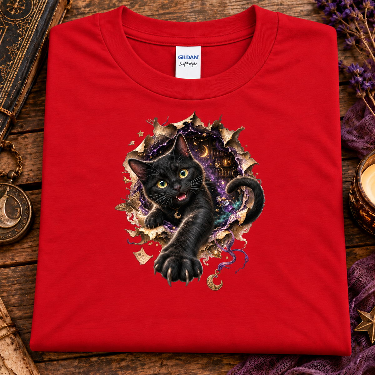 A folded red Gildan Softstyle t-shirt with a graphic of a black cat bursting through torn paper, displayed on a rustic wooden surface with decorative props.