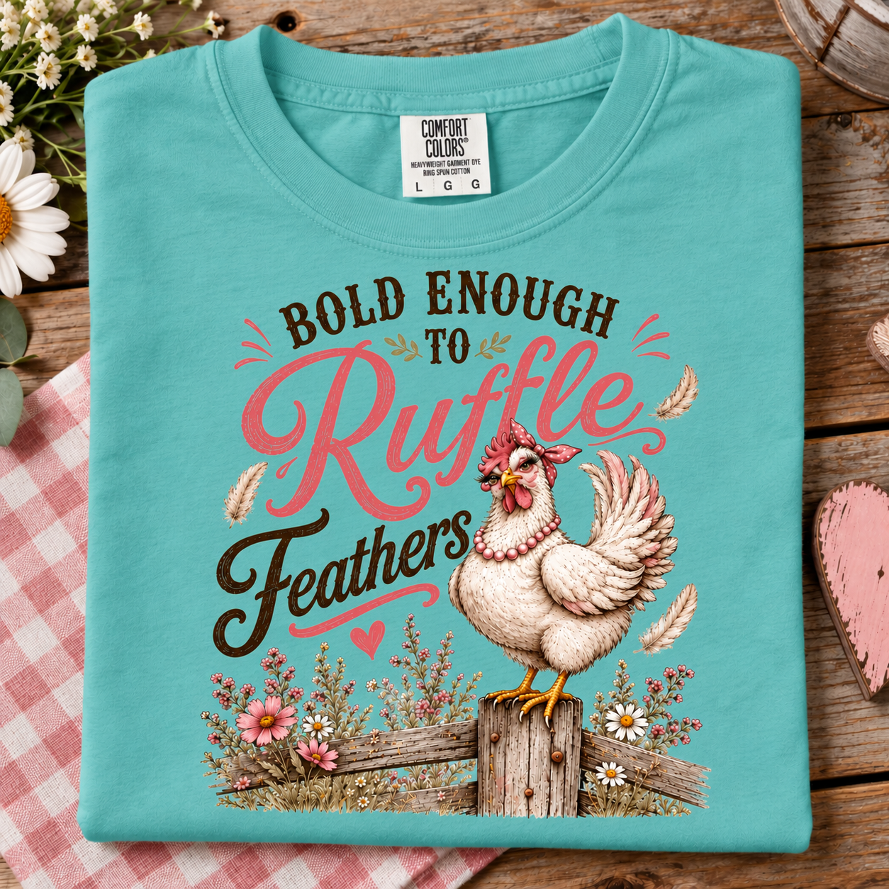 A folded chalky mint Comfort Colors t-shirt with a graphic print of a chicken and the text 'BOLD ENOUGH TO Ruffle Feathers', displayed on a rustic wooden background with a pink gingham cloth.