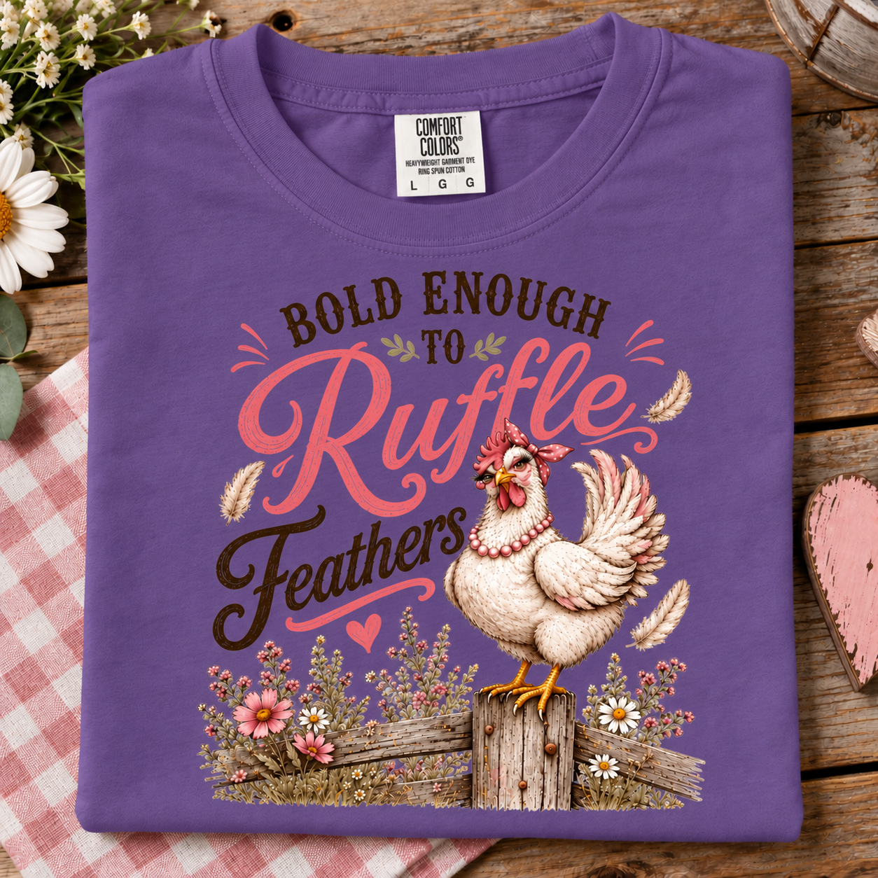 A folded purple t-shirt with a graphic print of a rooster and the text 'BOLD ENOUGH TO Ruffle Feathers' in pink, brown, and white, displayed on a rustic wooden background with a red gingham cloth and flowers.