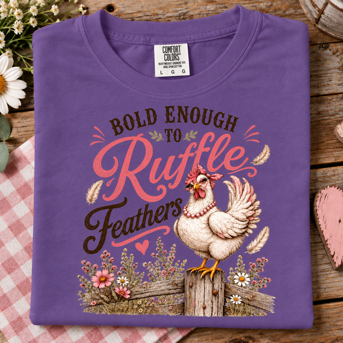 A folded purple t-shirt with a graphic print of a rooster and the text 'BOLD ENOUGH TO Ruffle Feathers' in pink, brown, and white, displayed on a rustic wooden background with a red gingham cloth and flowers.