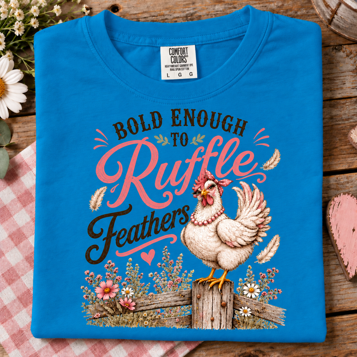 A folded sapphire colored Comfort Colors t-shirt with a graphic print of a chicken and the text 'BOLD ENOUGH TO Ruffle Feathers', displayed on a rustic wooden background with a pink gingham cloth.