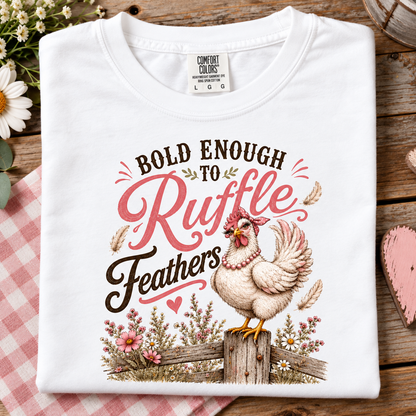 A folded white t-shirt with a graphic print of a chicken and the text 'BOLD ENOUGH TO Ruffle Feathers' in pink and brown lettering, displayed on a rustic wooden background with a pink gingham cloth and flowers.