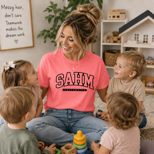 Woman in a pink 'SAHM University' shirt sitting with children in a home setting.