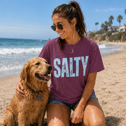 Woman wearing a berry 'SALTY' t-shirt sitting on the beach with a dog.
