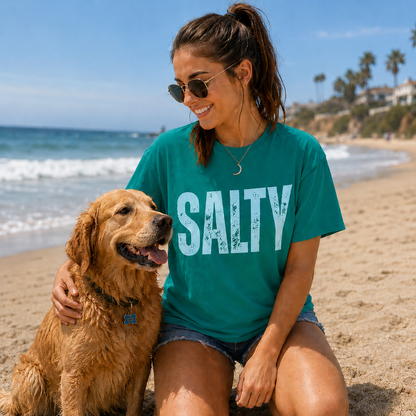 Woman wearing a Island Green 'SALTY' t-shirt sitting on the beach with a dog.