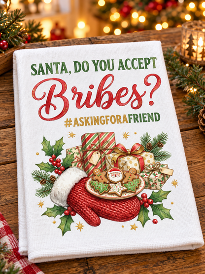 A white kitchen towel with a festive Christmas design, featuring the text 'SANTA, DO YOU ACCEPT Bribes? #ASKINGFORAFRIEND' above an illustration of a red mitten, gifts, holly, and gingerbread cookies, displayed on a wooden table with holiday lights in the background.