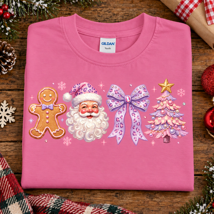 A folded azalea pink Gildan t-shirt with a Christmas-themed graphic print featuring a gingerbread man, Santa Claus, a purple bow, and a decorated Christmas tree, displayed on a rustic wooden background with holiday decorations.