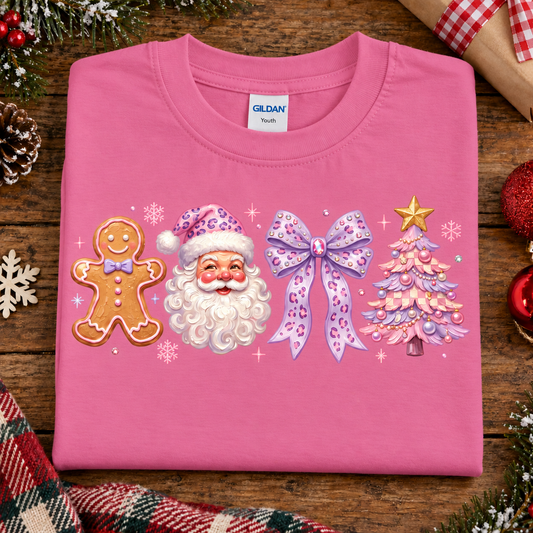 A folded azalea pink Gildan t-shirt with a Christmas-themed graphic print featuring a gingerbread man, Santa Claus, a purple bow, and a decorated Christmas tree, displayed on a rustic wooden background with holiday decorations.
