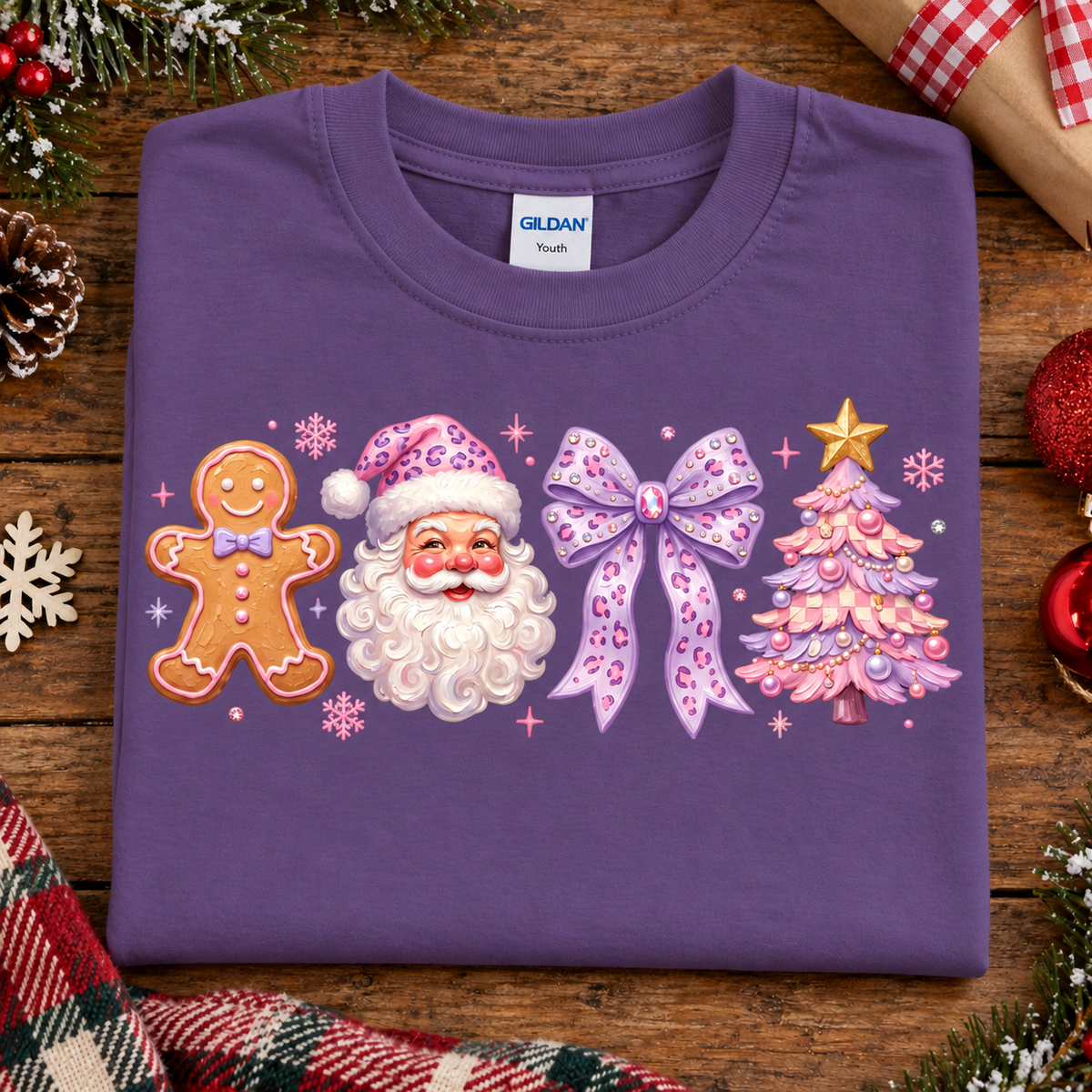 A purple Gildan Youth t-shirt with a Christmas-themed graphic print featuring a gingerbread man, Santa Claus, a purple bow, and a pink Christmas tree, laid flat on a rustic wooden table surrounded by holiday decorations.