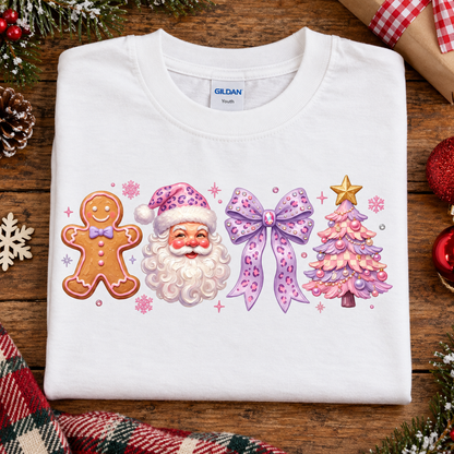 A folded white Gildan T-shirt laid flat on a rustic wooden surface, decorated with a colorful Christmas-themed graphic featuring a gingerbread man, Santa Claus, a pink bow, and a pink Christmas tree. The shirt is surrounded by holiday decorations including pine branches, a red plaid scarf, and ornaments.