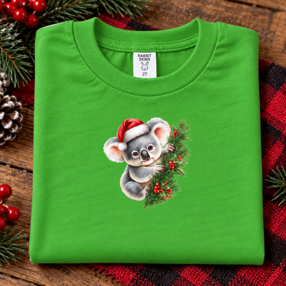 A folded apple green t-shirt with a cartoon koala wearing a Santa hat and holding a Christmas wreath, displayed on a rustic wooden background with holiday decorations.