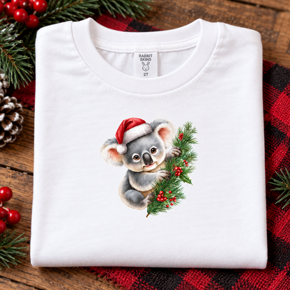 A folded white crewneck t-shirt featuring a printed illustration of a cute gray koala wearing a red Santa hat and holding a green Christmas branch with red berries. The shirt is styled on a rustic wooden surface with a red and black plaid blanket, pine branches, and red berries.