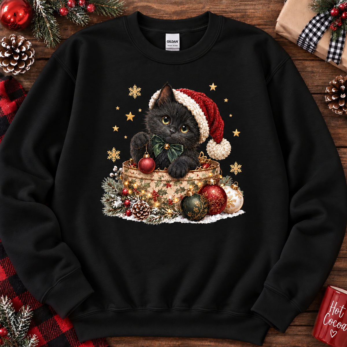 A black crewneck sweatshirt featuring a festive Christmas graphic of a black kitten wearing a red Santa hat and bow tie, sitting in a gift box surrounded by ornaments and pinecones. The sweatshirt is displayed flat on a rustic wooden background decorated with holiday items like pinecones, a wrapped gift, and red berries.