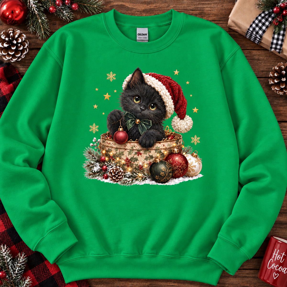 A irish green crewneck sweatshirt featuring a festive Christmas graphic of a black cat in a Santa hat sitting in a basket with ornaments. The sweatshirt is displayed in a flat-lay arrangement on a rustic wooden background with holiday decorations like pine cones, a wrapped gift, and red ornaments.