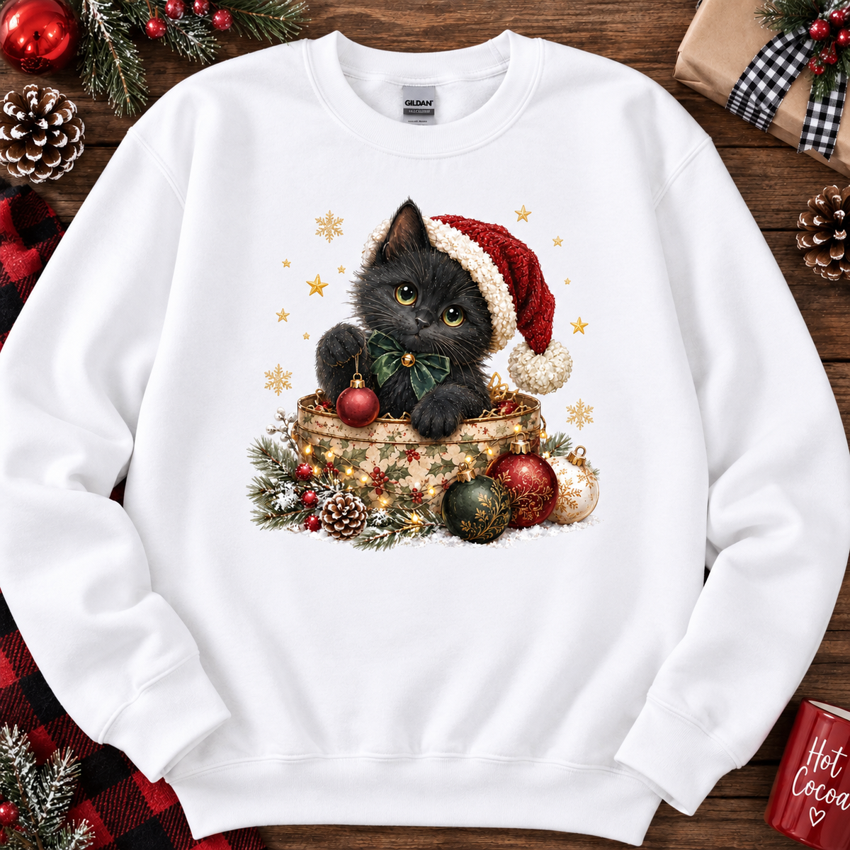 A white crewneck sweatshirt featuring a festive graphic of a black kitten wearing a red Santa hat and bow, sitting in a decorated gift box. The sweatshirt is displayed on a rustic wooden background surrounded by holiday decorations like pine cones, ornaments, and a plaid blanket.