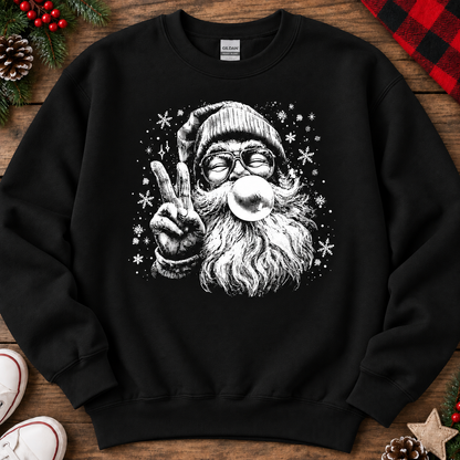 A black crewneck sweatshirt featuring a white graphic of a festive Santa Claus making a peace sign while blowing a bubblegum bubble, surrounded by snowflakes. The sweatshirt is displayed in a flat-lay arrangement on a rustic wooden background with holiday decorations including pine branches, red berries, a red and black plaid blanket, and a white sneaker.
