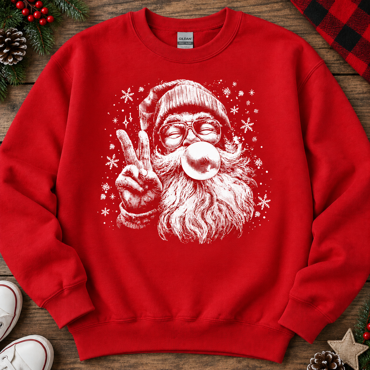 A red crewneck sweatshirt featuring a white graphic of a Santa Claus character blowing a bubblegum bubble and making a peace sign, laid flat on a rustic wooden surface with holiday decorations.