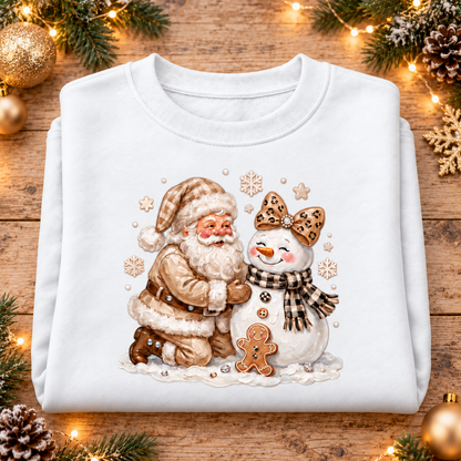 A folded white crewneck sweatshirt with a festive Christmas graphic of Santa Claus kneeling next to a snowman, displayed on a rustic wooden background decorated with pine branches, fairy lights, and ornaments.