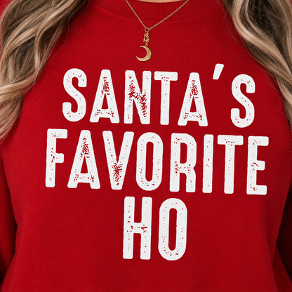 A close-up of a red crewneck sweatshirt with the text 'SANTA'S FAVORITE HO' printed in distressed white letters. A model's blonde hair, neck, and a gold crescent moon pendant necklace are visible.
