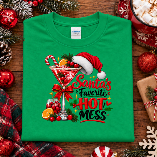 A festive Christmas-themed green colored graphic t-shirt featuring a martini glass filled with red holiday drink, candy cane, cranberries, orange slices, pine sprigs, a red bow, a Santa hat, and the text 'Santa's Favorite HOT MESS' on a white background.