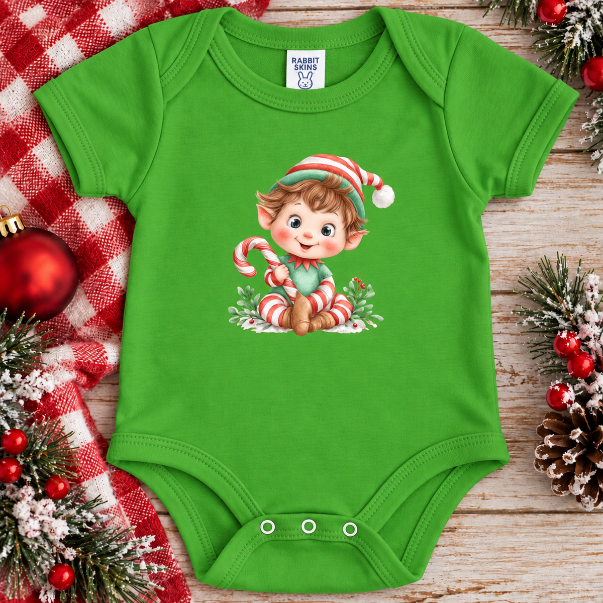 A apple green baby bodysuit with a cartoon elf holding a candy cane printed on the chest, styled in a flat lay on a rustic wooden surface with red-and-white check fabric, ornaments, pinecones, and holiday greenery.