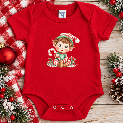 A baby red short-sleeve bodysuit with a festive Christmas elf graphic on the chest, styled in a flat lay on a rustic wooden surface with holiday decorations including a red and white checkered blanket, pine branches, and ornaments.