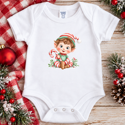 A white baby bodysuit with a festive Christmas elf graphic, styled in a flat lay on a wooden surface with holiday decorations including a red plaid blanket, pine branches, and ornaments. 