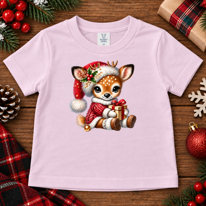 A light pink toddler t-shirt featuring a cute Christmas deer graphic, styled in a flat-lay on a rustic wooden background with holiday decorations like pine branches, a red ornament, and a plaid scarf.